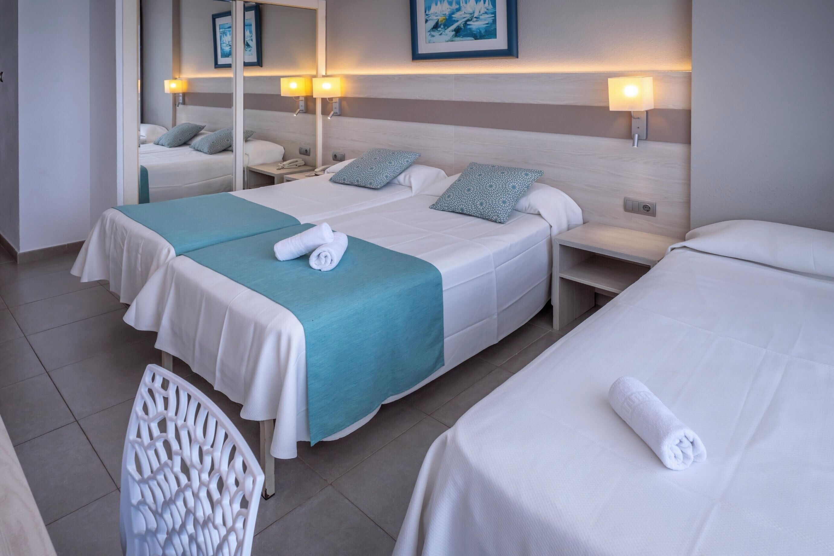 A hotel room with two beds, each with a white bedspread and a teal blanket. There are two bedside tables with lamps, a mirror, and a framed picture on the wall.