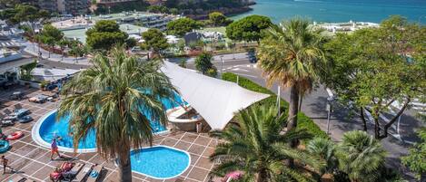 A pool area with lounge chairs, palm trees, and a view of the sea and buildings in the background.
