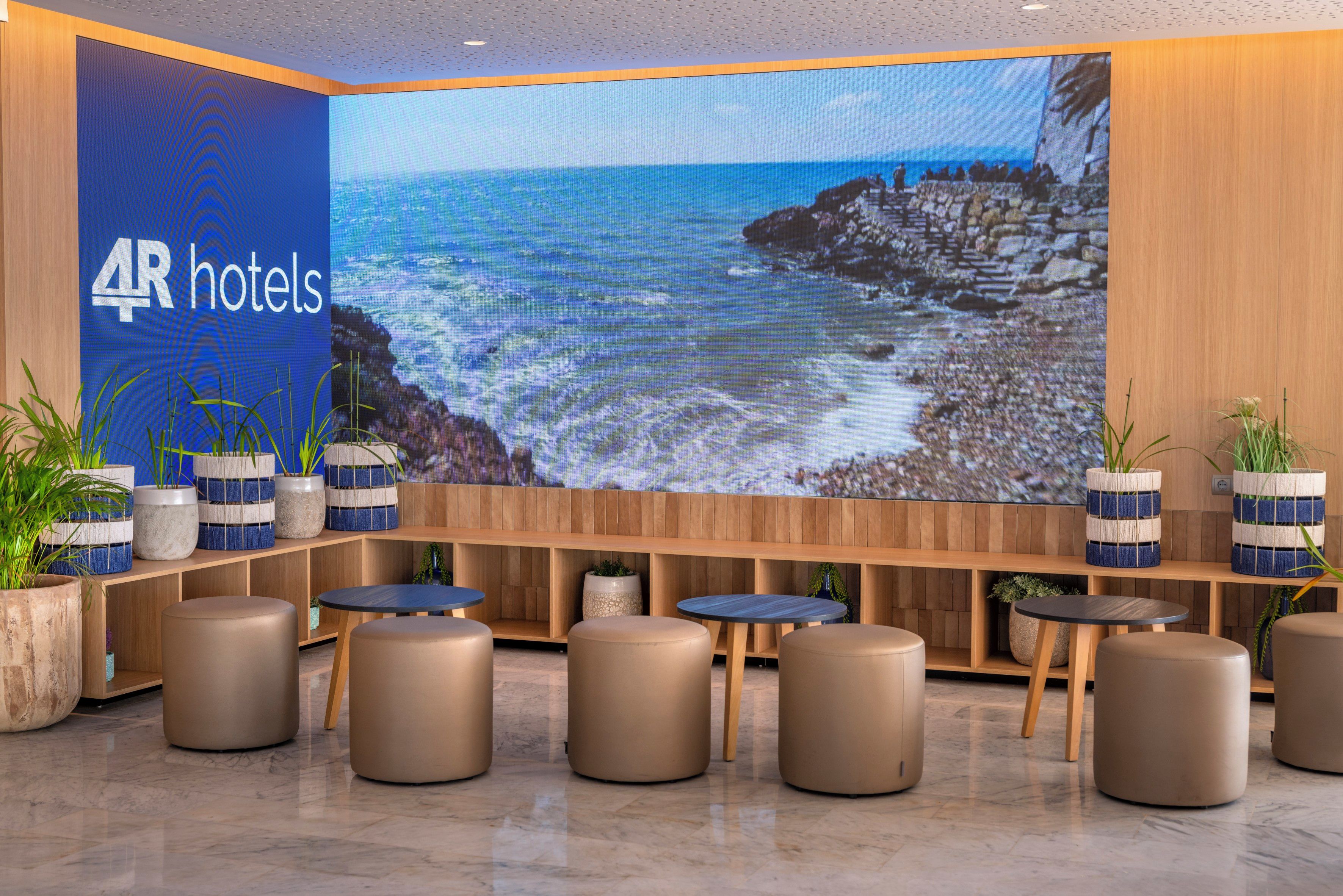 Um hall de hotel com uma tela digital grande exibindo uma paisagem costeira, áreas de estar com mesas redondas e pufes, e plantas em vasos.