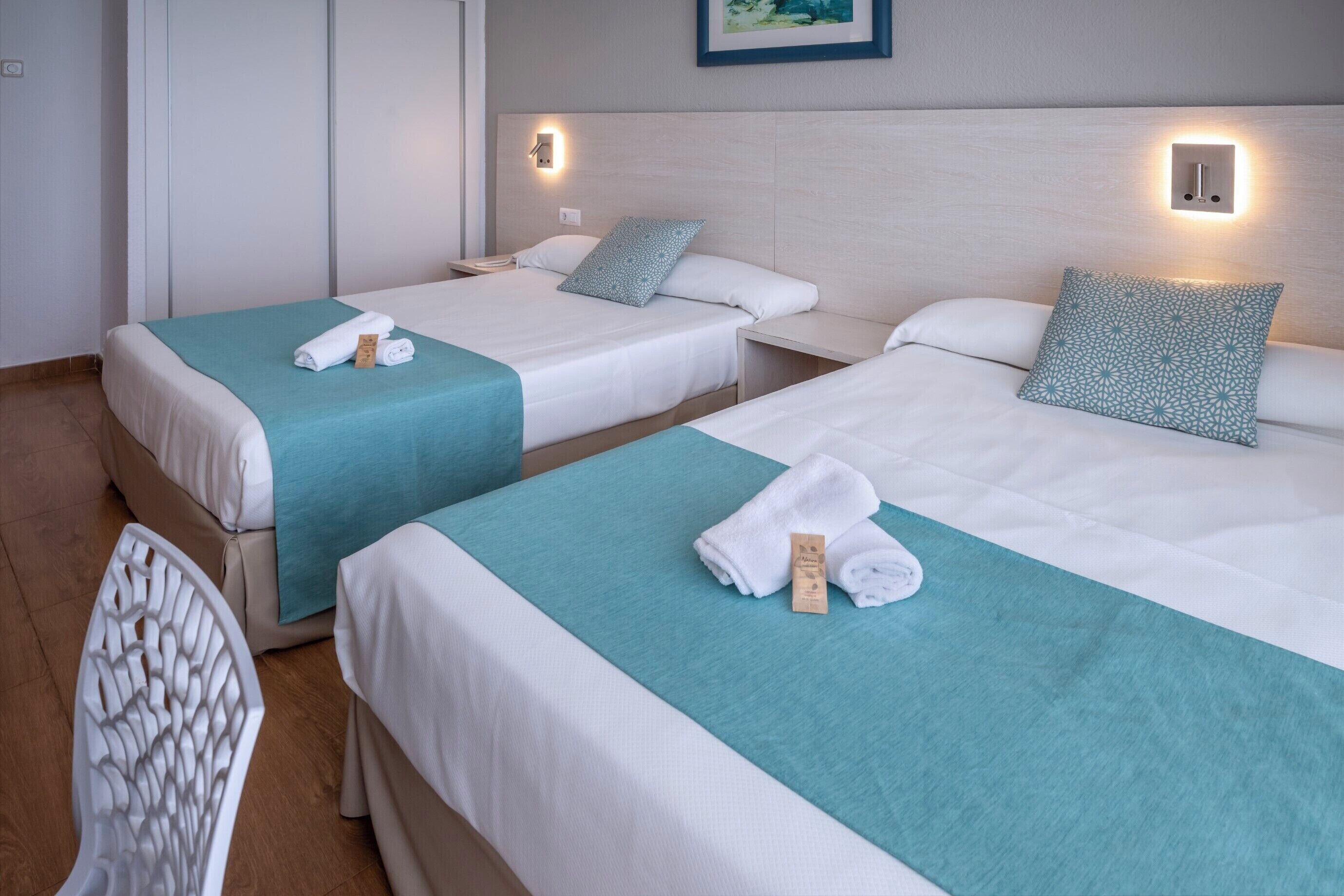 Une chambre d’hôtel avec deux lits, chacun recouvert d’une couvre-lit turquoise et doté de draps blancs. Des lampes murales sont fixées au-dessus des lits, et une petite table se trouve entre les deux.