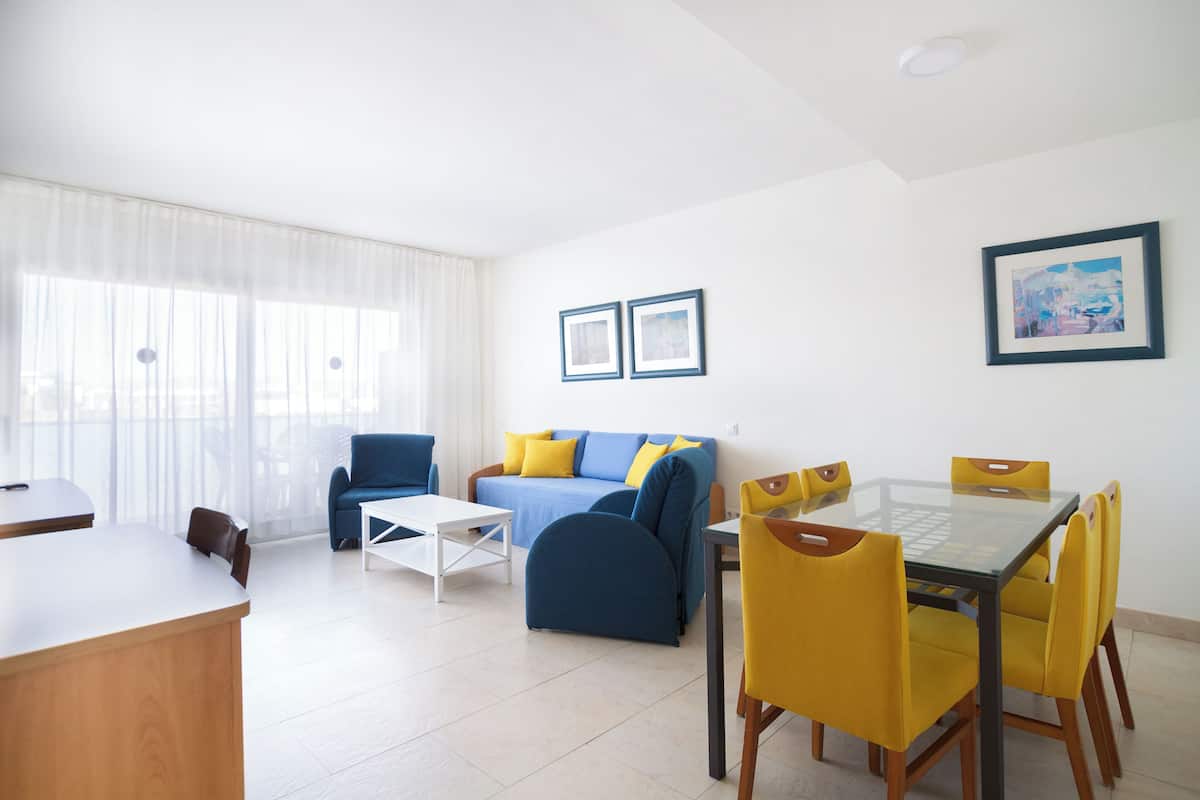 A modern living room with a sofa, coffee table, and dining table with yellow chairs. There are two framed pictures on the wall and a window with sheer curtains.