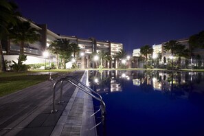 A nighttime view of a swimming pool with a metal ladder, surrounded by palm trees and modern buildings with illuminated windows. - Ibersol Spa Aqquaria (Salou)