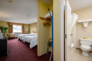 A hotel room with two beds, a desk, a chair, a plant, a shower, and a toilet.
