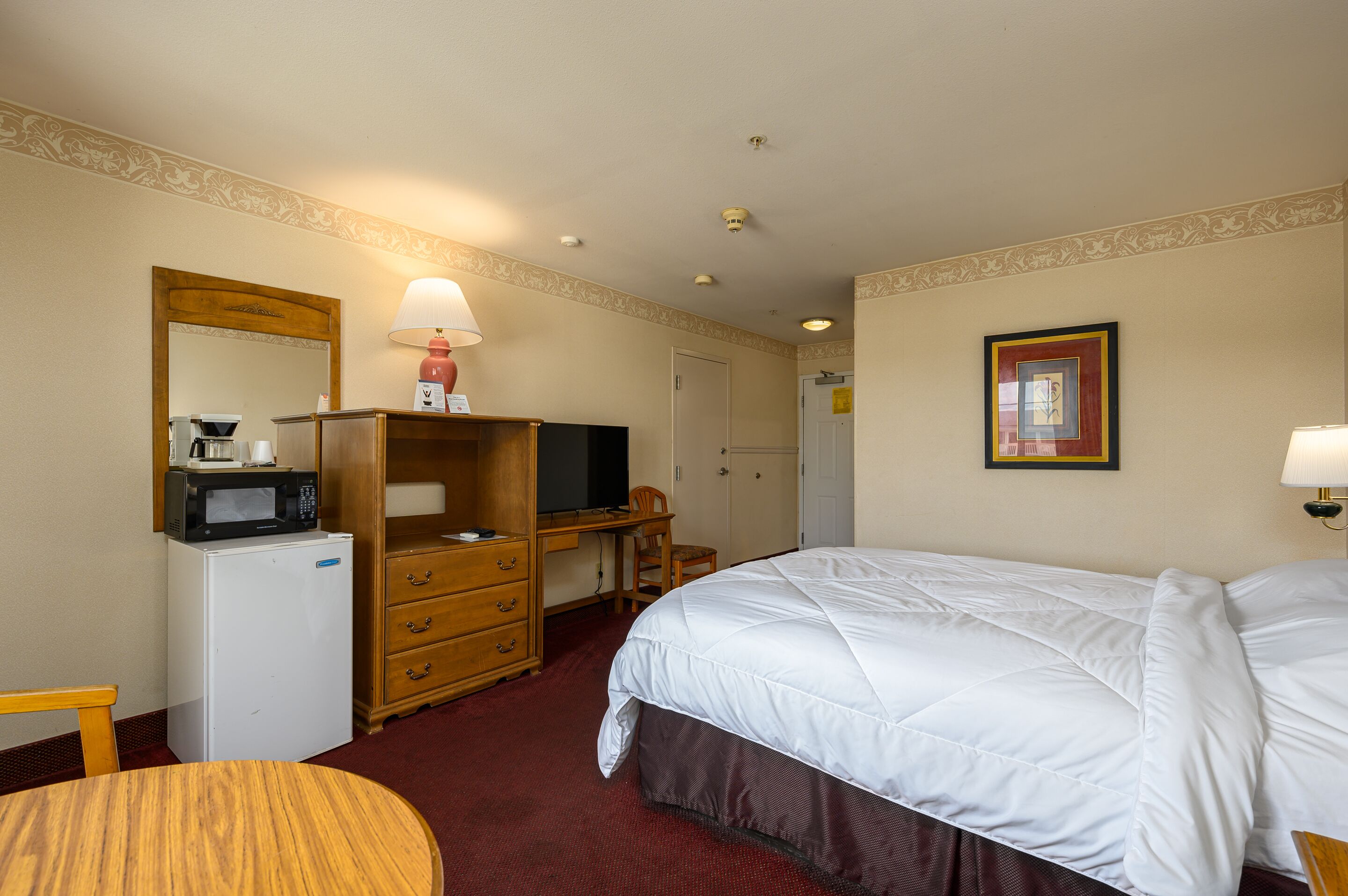 A hotel room with a bed, a desk with a computer, a refrigerator, a microwave, a chair, and a small table.