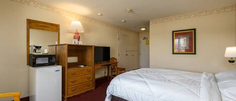 A hotel room with a bed, a desk with a computer, a refrigerator, a microwave, a chair, and a small table.