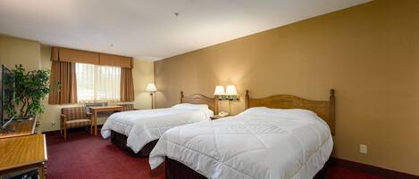 A hotel room with two beds, a desk, a chair, a lamp, and a plant.