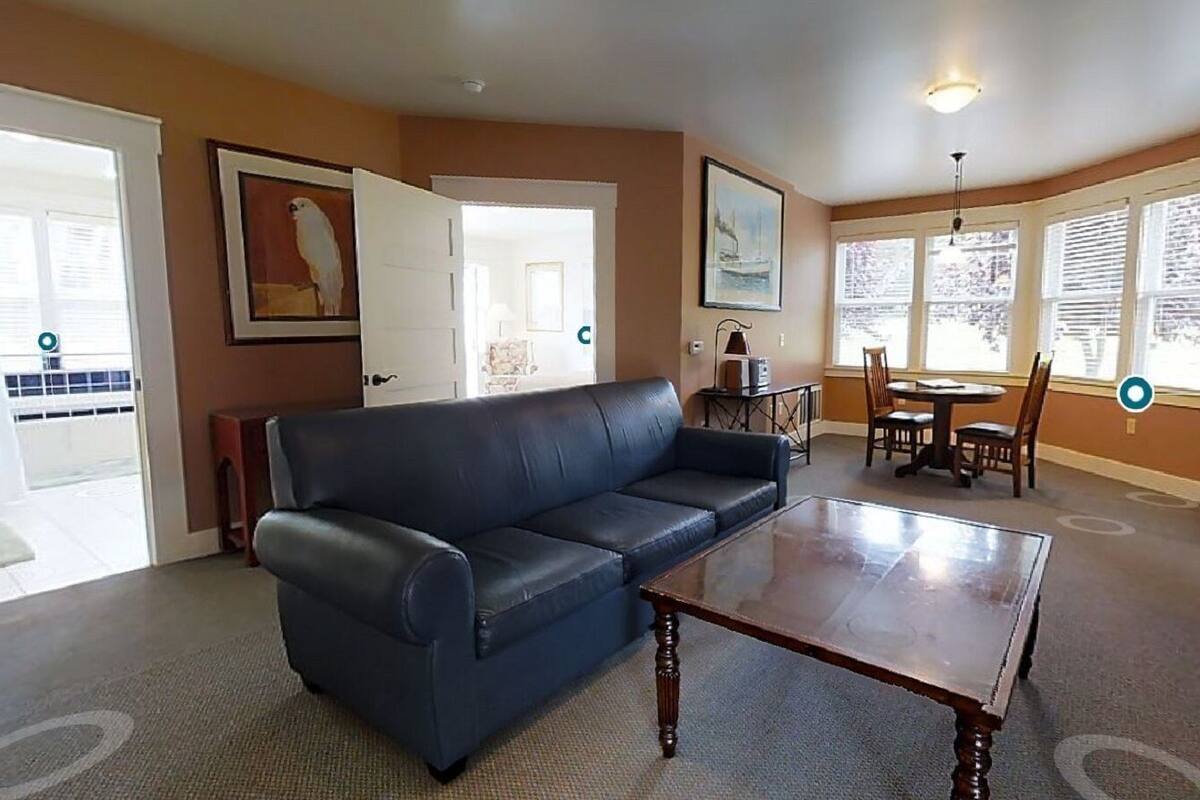 A living room with a dark leather sofa, a wooden coffee table, and a fireplace. There are framed pictures on the walls and a dining area with a table and chairs.