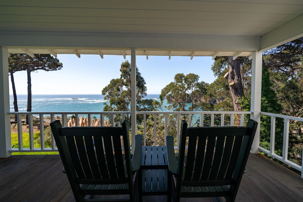 A covered outdoor area with two wooden chairs and a table, overlooking a coastal view with trees and the ocean.