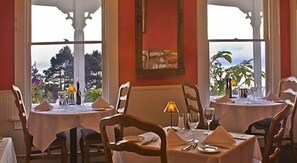 A dining area with round tables, chairs, and a view of trees through large windows.
