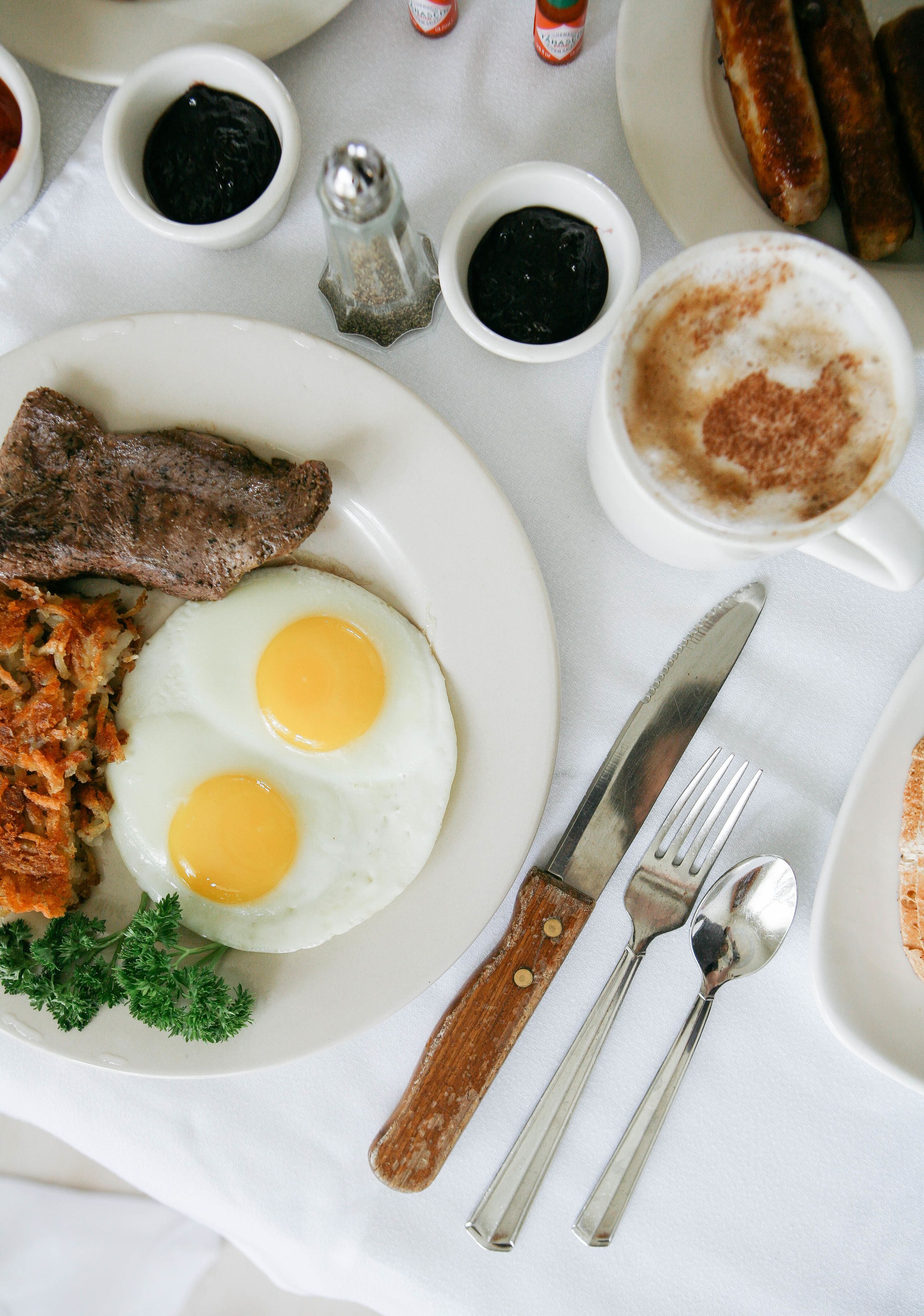 Prato de pequeno-almoço com ovos, bife e batata palha, acompanhado de acompanhamentos e talheres sobre uma mesa.
