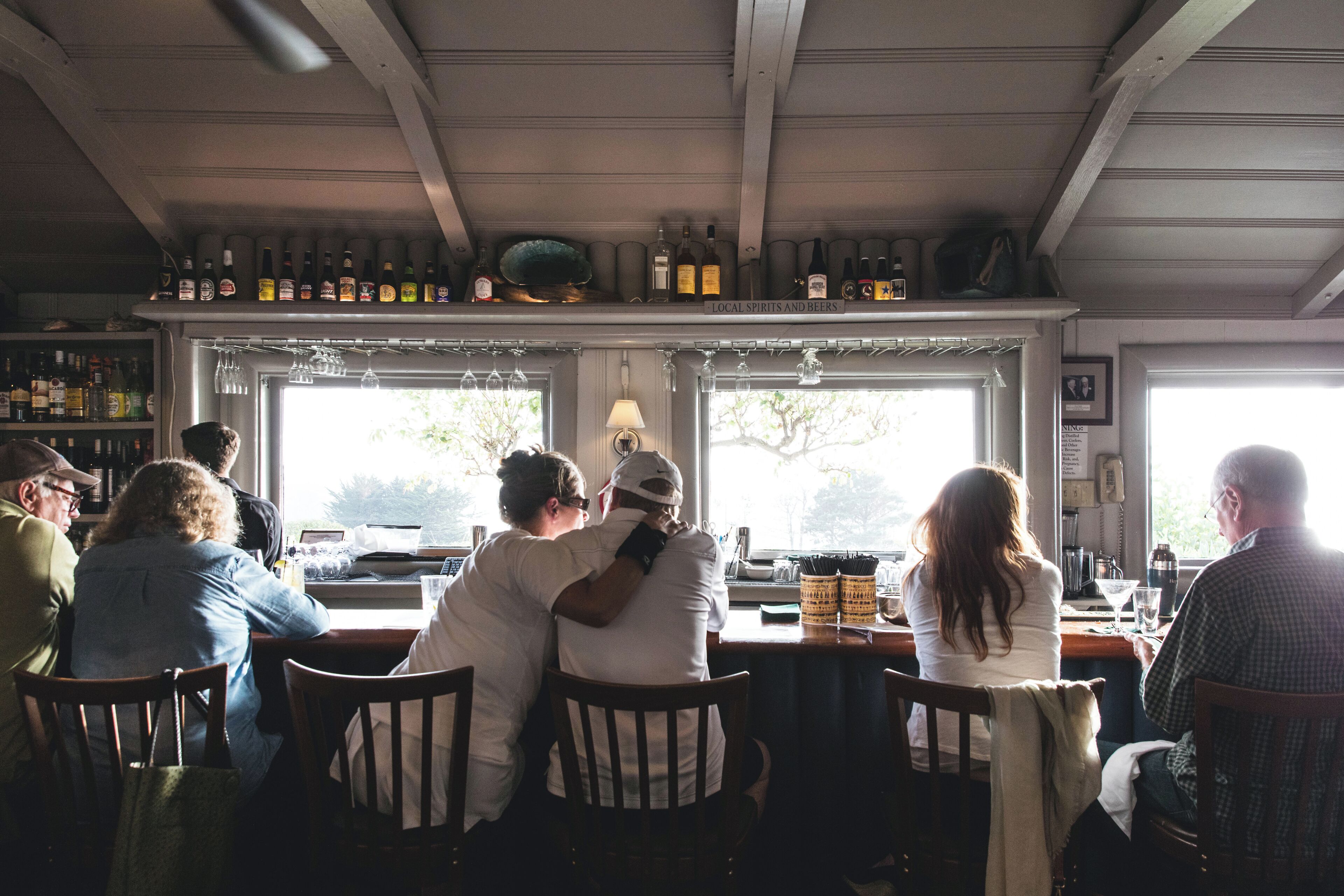 Un espace bar avec des clients assis à des tables, une variété de bouteilles exposées sur des étagères et une fenêtre donnant sur l’extérieur.