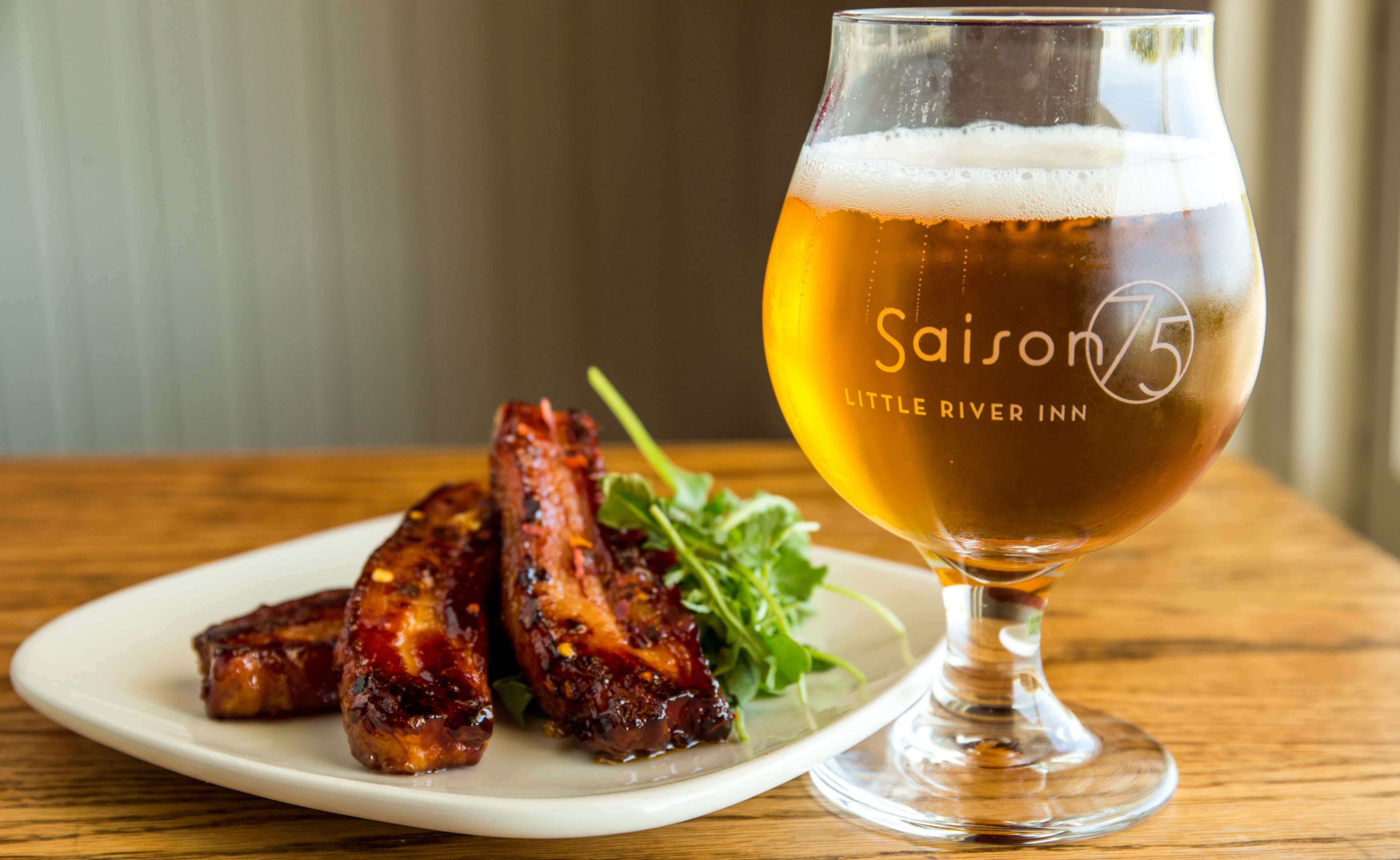 A glass of beer with "Saison 75" and "LITTLE RIVER INN" on it, alongside a plate of barbecued ribs and greens.