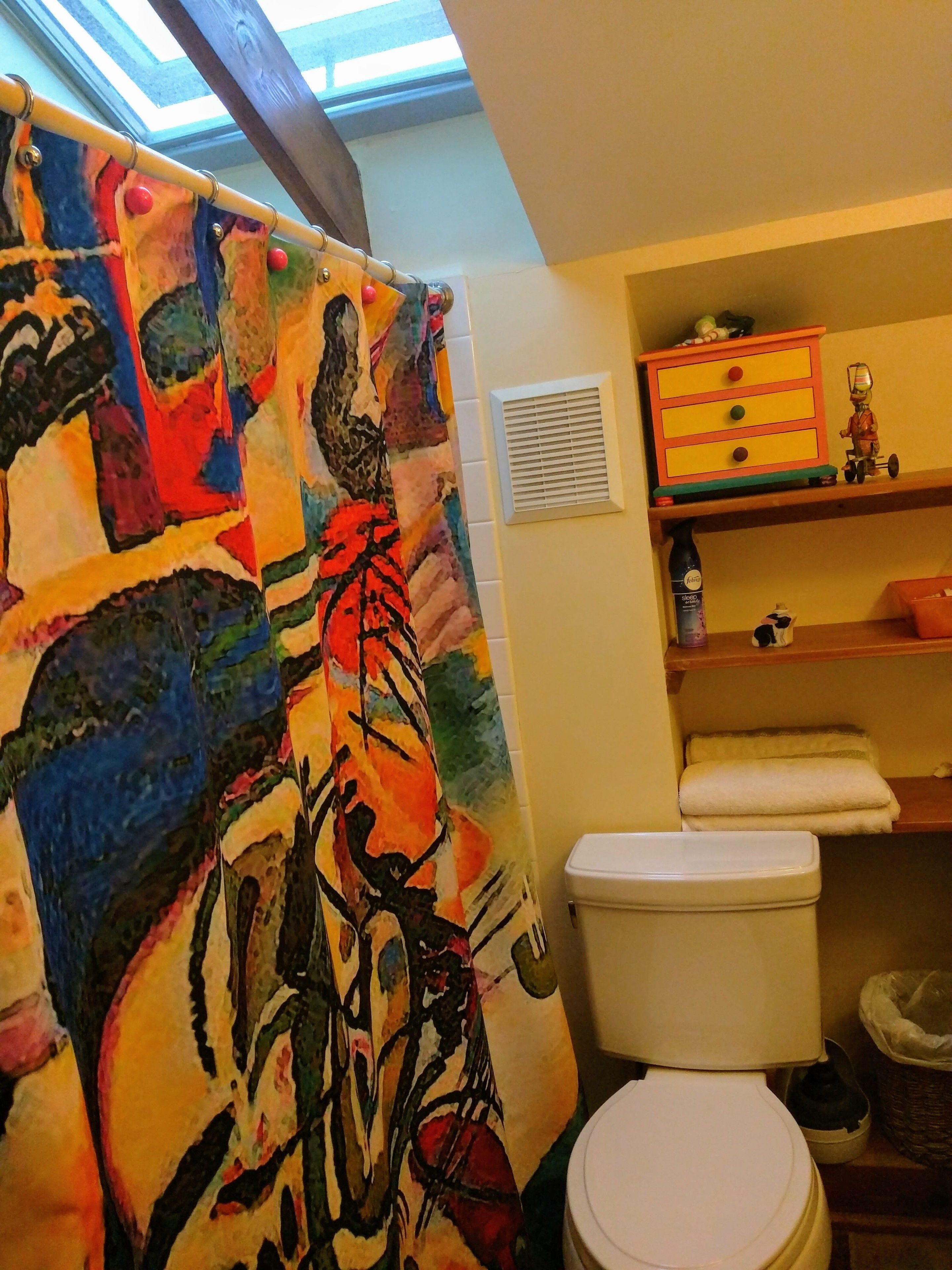 Un baño con una cortina de ducha colorida, un inodoro, un estante con juguetes y una ventana sobre la ducha.