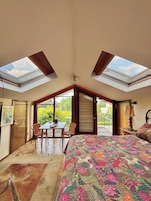 Un dormitorio con tragaluz, una cama, una mesa con sillas y vistas al verde exterior.