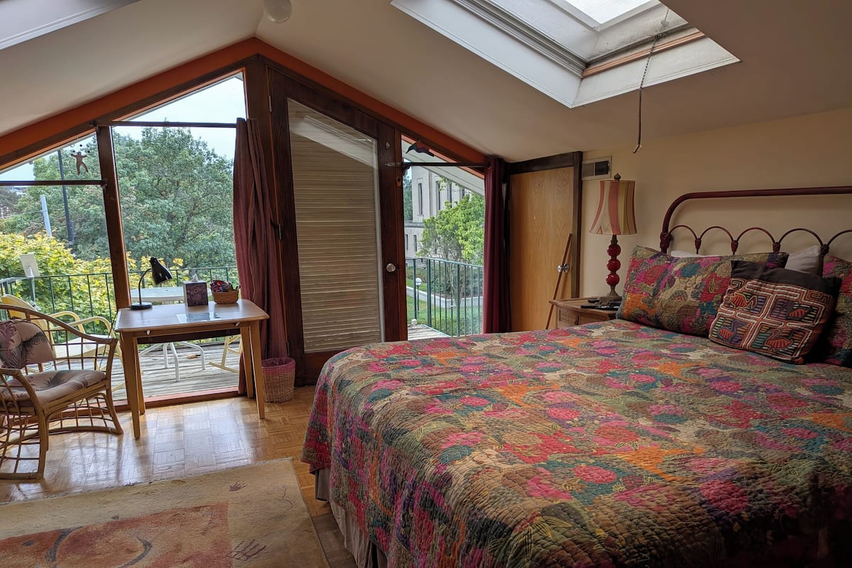 A bedroom with a large bed, a desk, and a chair. There is a skylight above the bed and a view of the outdoors.