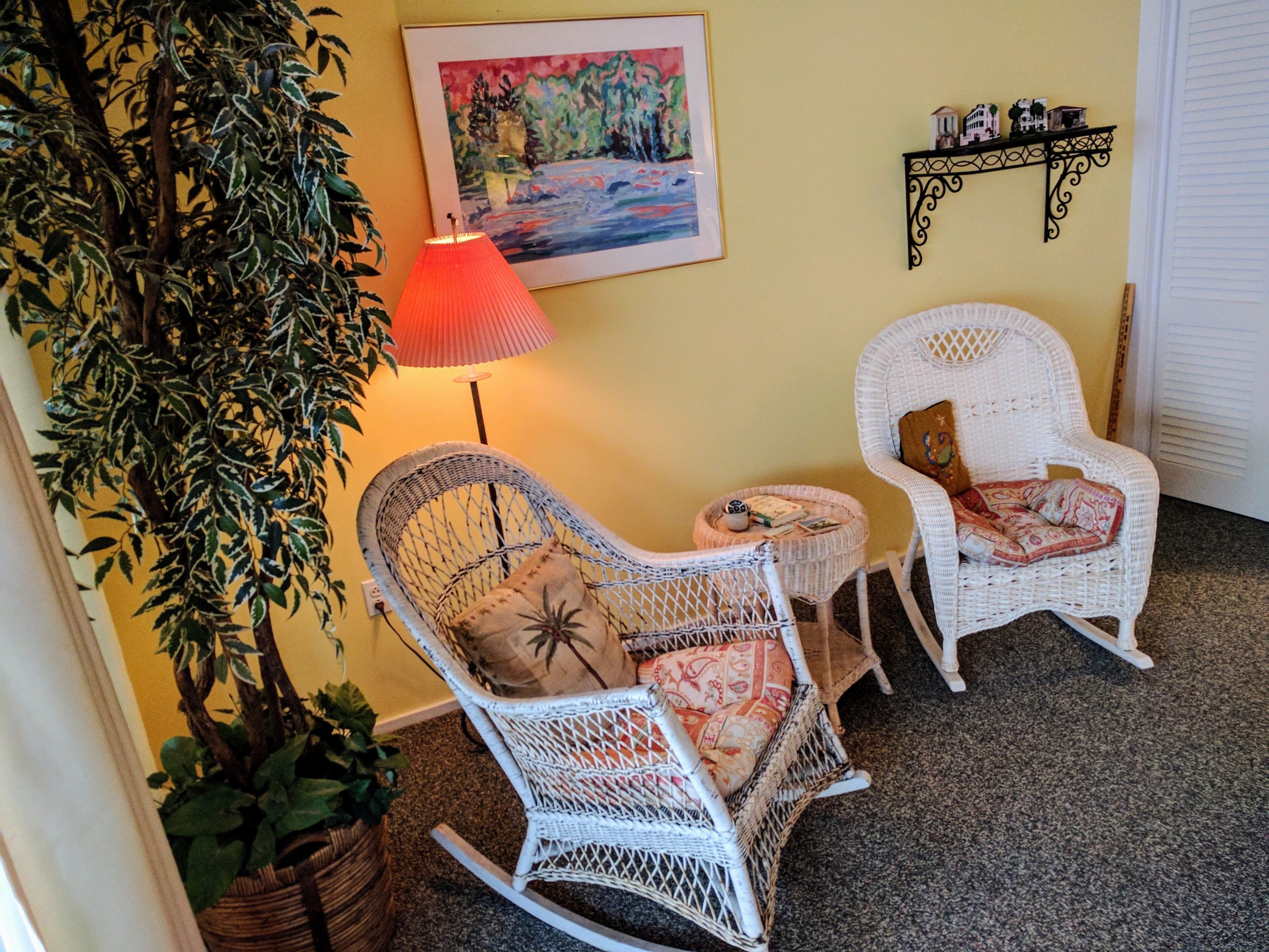 A room with two wicker rocking chairs, a small side table, a floor lamp, a potted plant, and a framed painting on the wall.