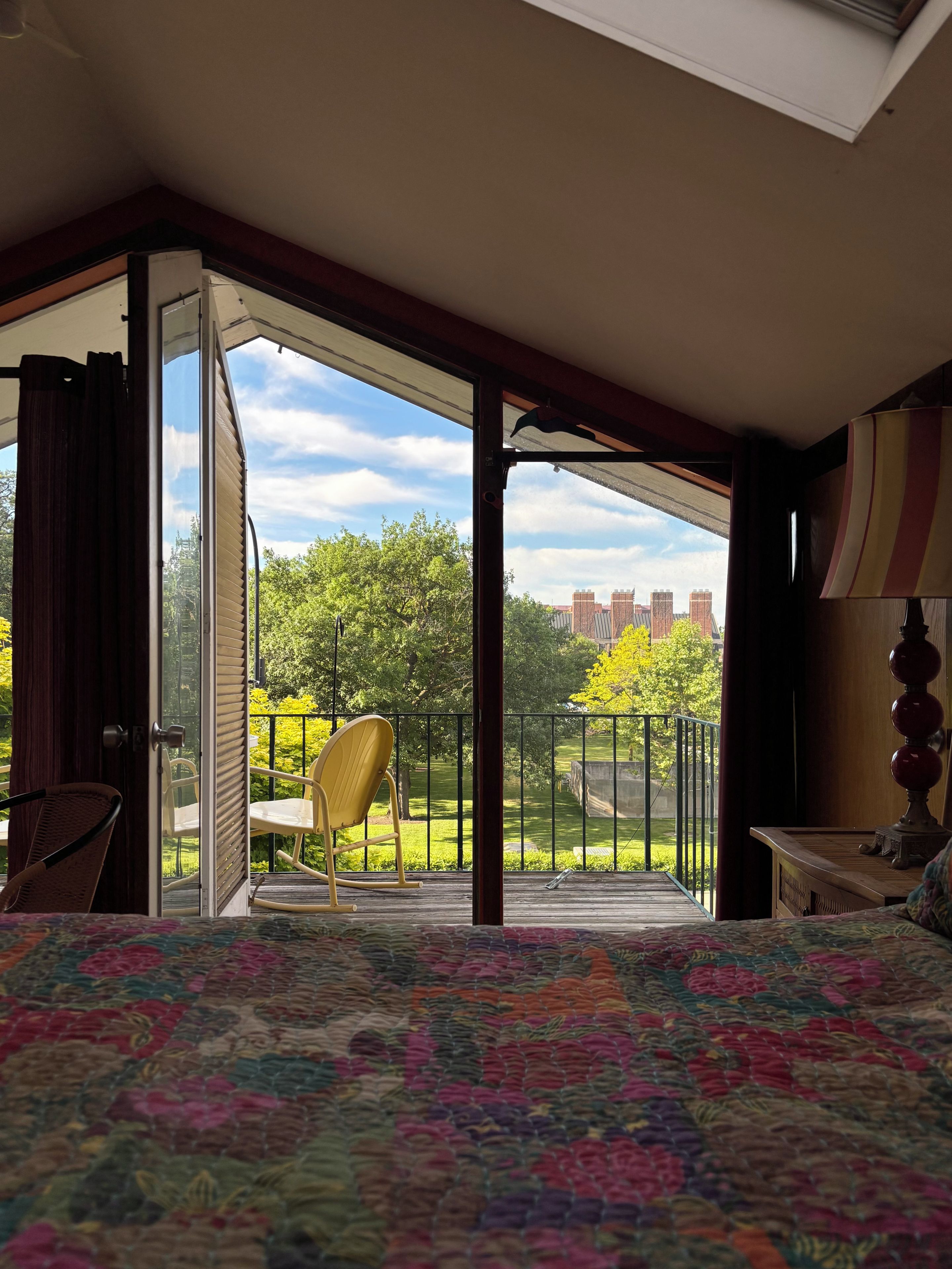Une chambre avec un grand lit, une chaise et une vue sur un parc et des immeubles, visible à travers une porte ouverte.