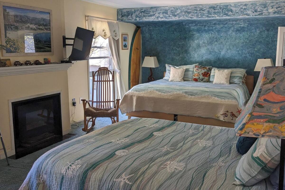 A bedroom with a fireplace, two beds, a chair, and a large painting.