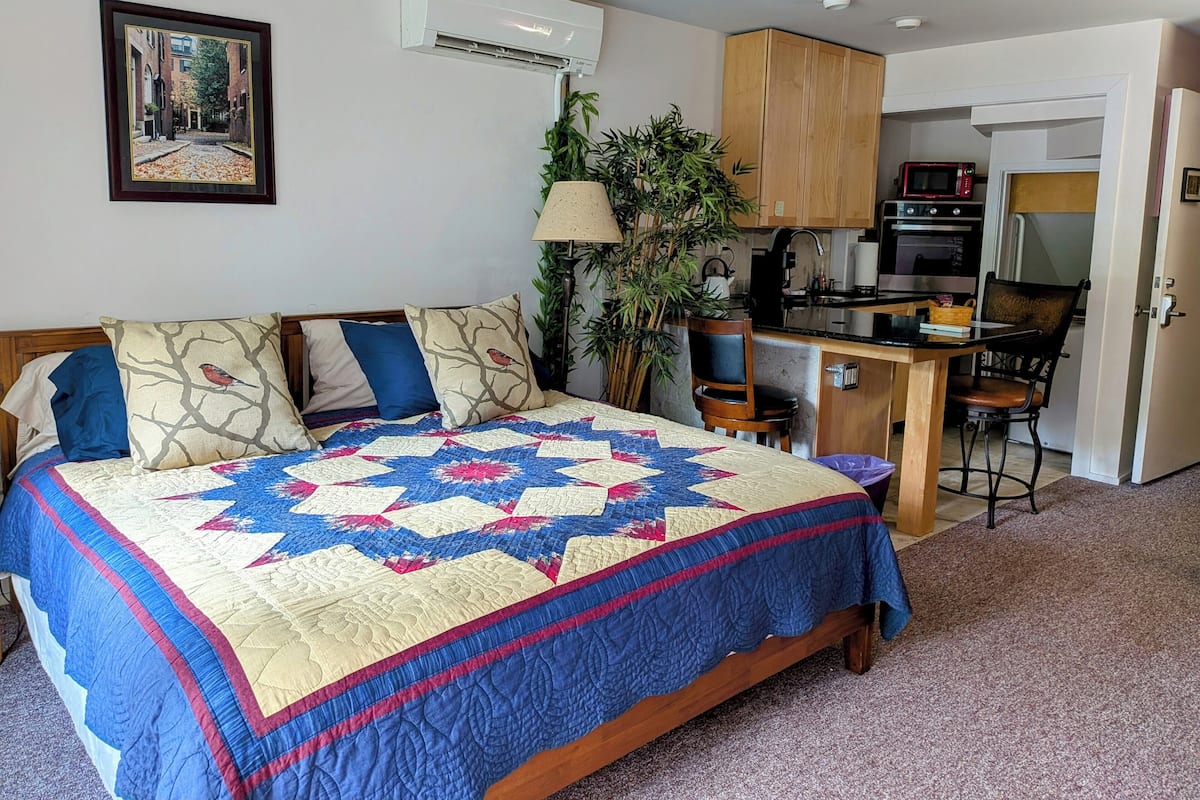 A bedroom with a large bed, a desk, a chair, an air conditioner, a microwave, and a door leading to a kitchen.