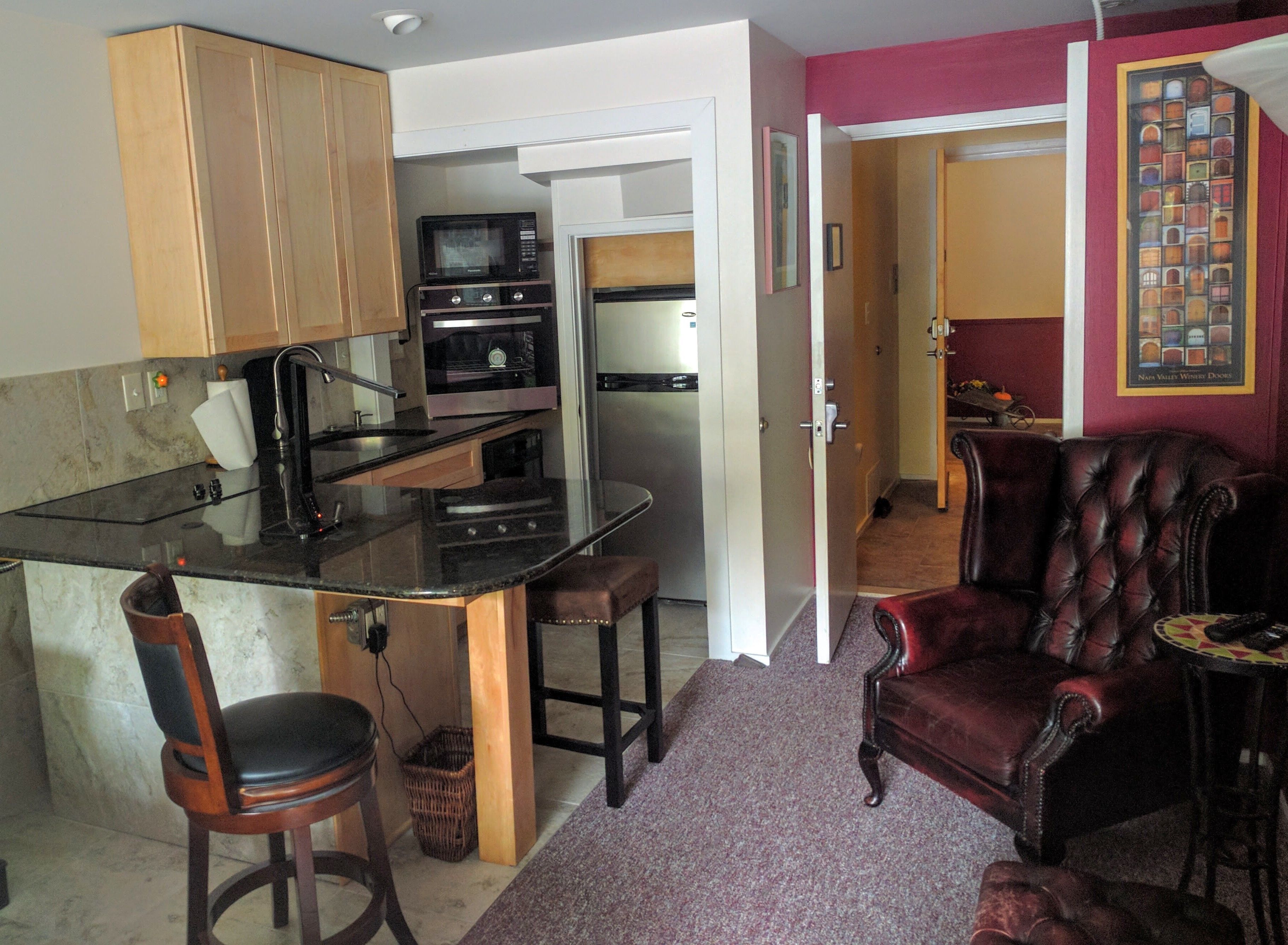 A compact kitchen with a microwave, oven, and sink. There is a dining area with a chair and a small table. A leather armchair and a framed picture are visible in the living room.