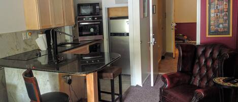 A compact kitchen with a microwave, oven, and sink. There is a dining area with a chair and a small table. A leather armchair and a framed picture are visible in the living room.