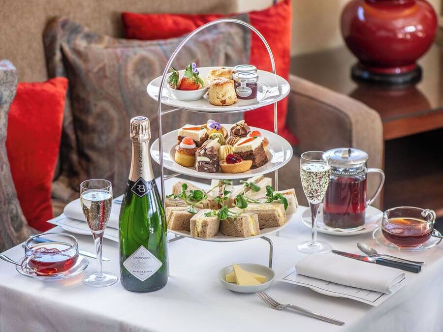 Ein dreistöckiges Dessertregal mit verschiedenen Gebäcken und Sandwiches, eine Champagnerflasche und eine Teekanne auf einem Tisch, der für eine Mahlzeit gedeckt ist.