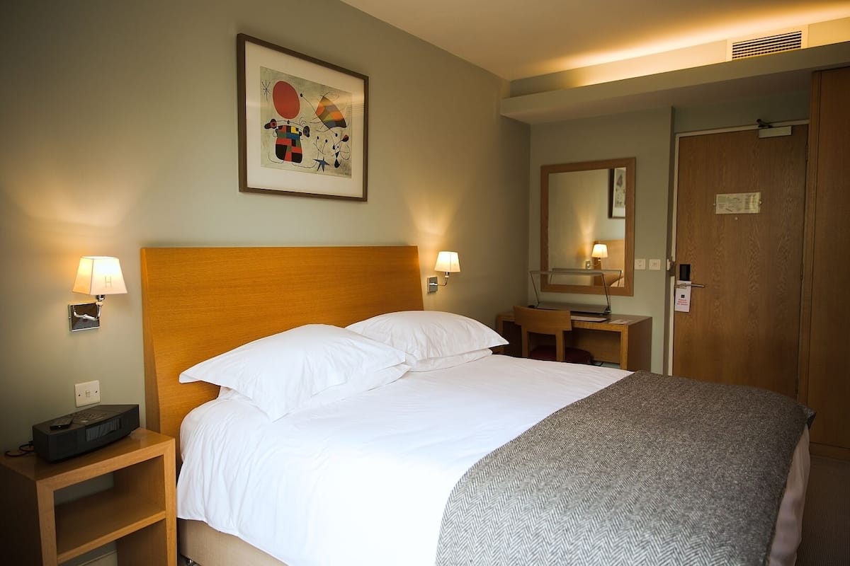A hotel room with a large bed, two bedside tables, a desk, a chair, a mirror, and a framed artwork on the wall.