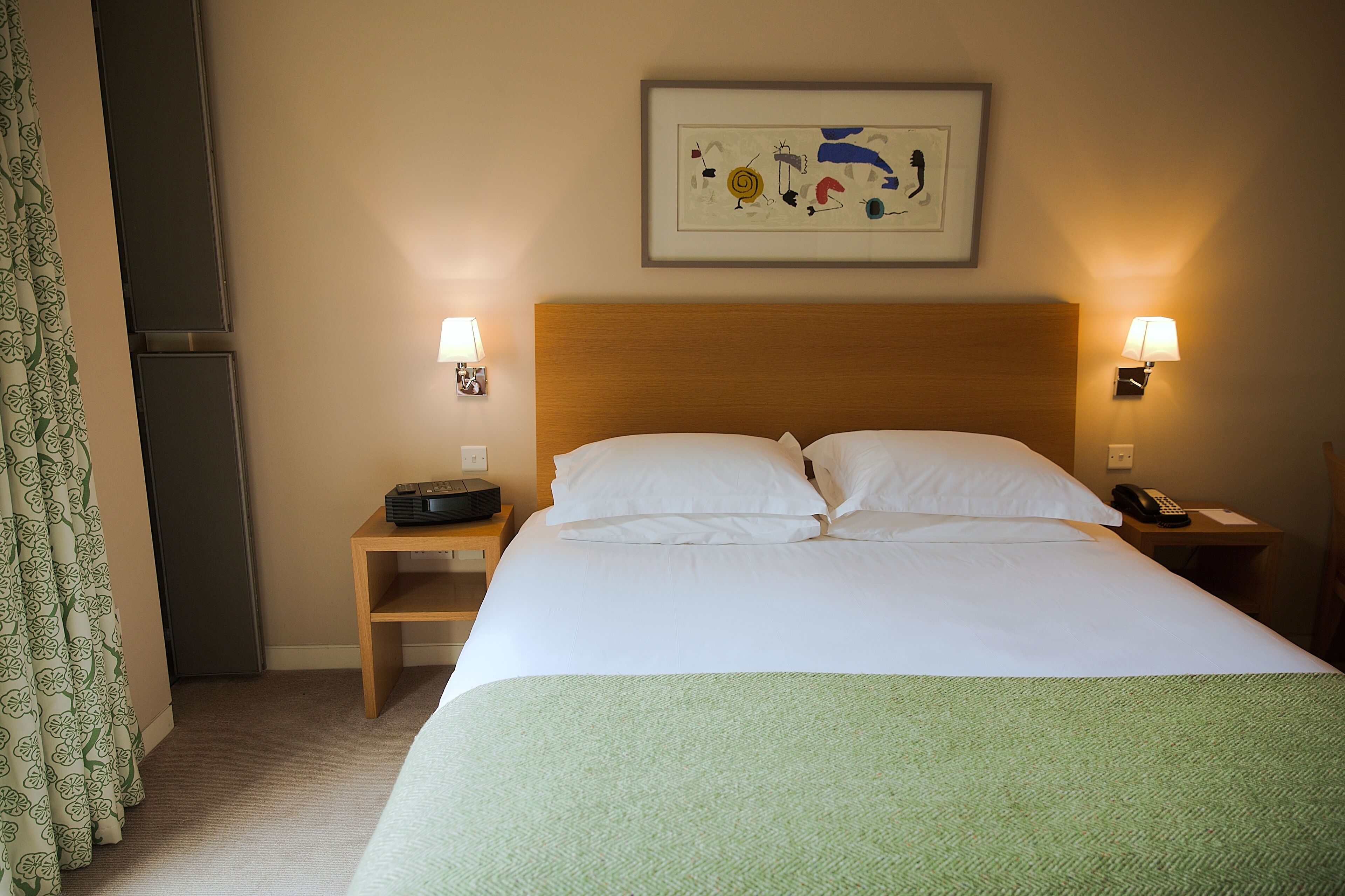 A hotel room with a bed, two bedside tables, a wall-mounted lamp, a framed artwork, and a curtain.