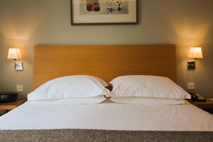 Una cama bien hecha con sábanas blancas, dos lámparas de noche y una obra enmarcada en la pared.