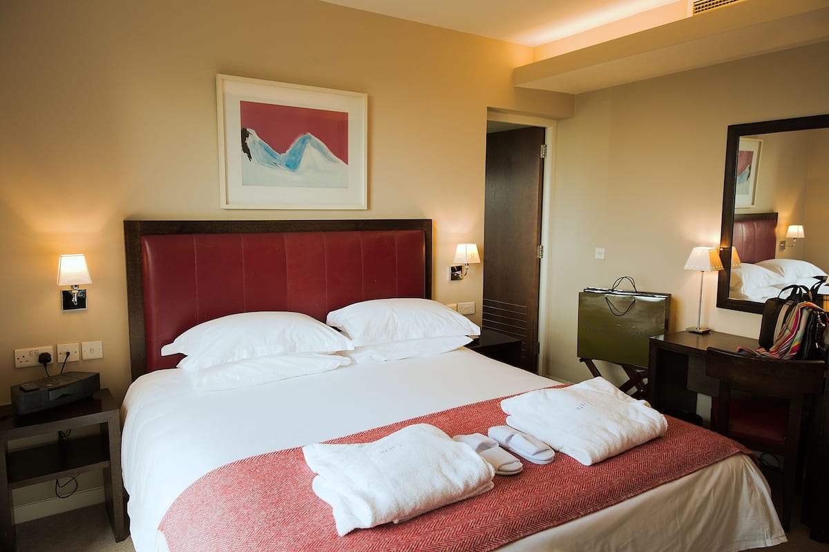 A hotel room with a large bed, a red headboard, white bedding, a red and white patterned throw, a nightstand with a lamp, a desk with a mirror, and a framed artwork on the wall.