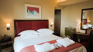 A hotel room with a large bed, a red headboard, white bedding, a red and white patterned throw, a nightstand with a lamp, a desk with a mirror, and a framed artwork on the wall.
