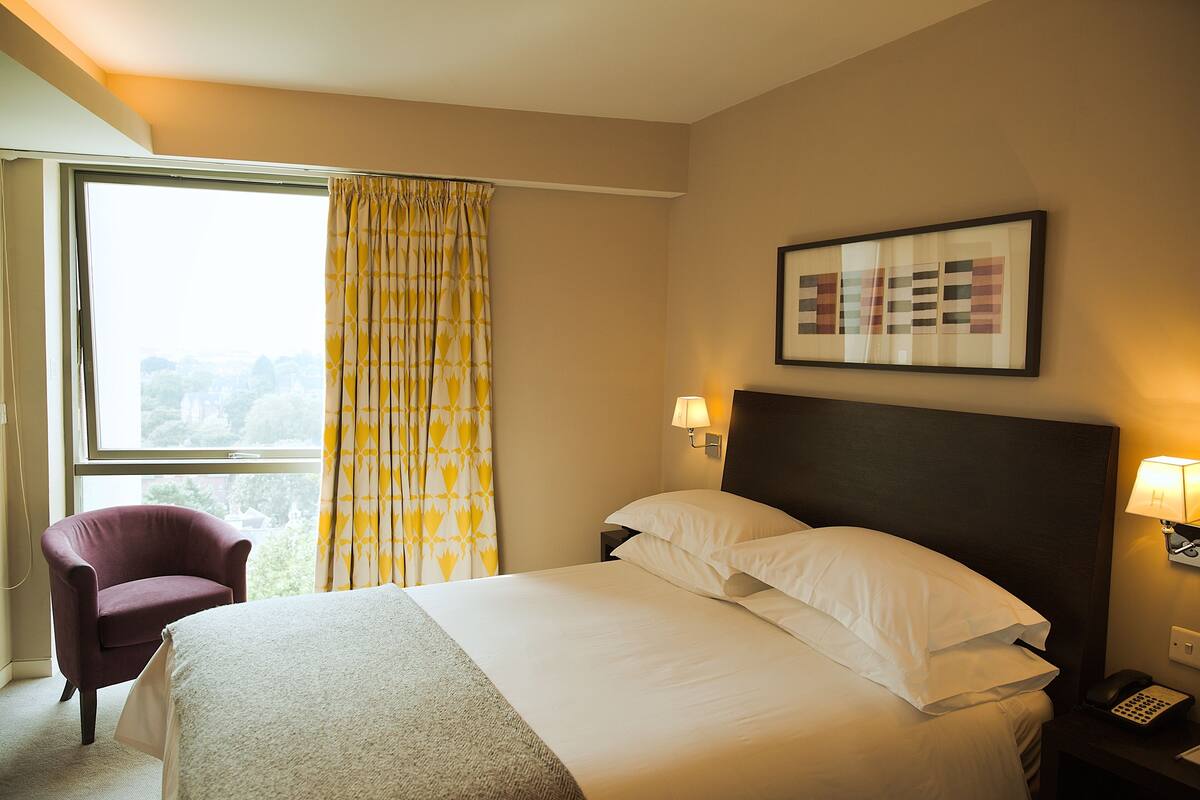A hotel room with a bed, a chair, a window with curtains, a wall-mounted lamp, and a framed picture.