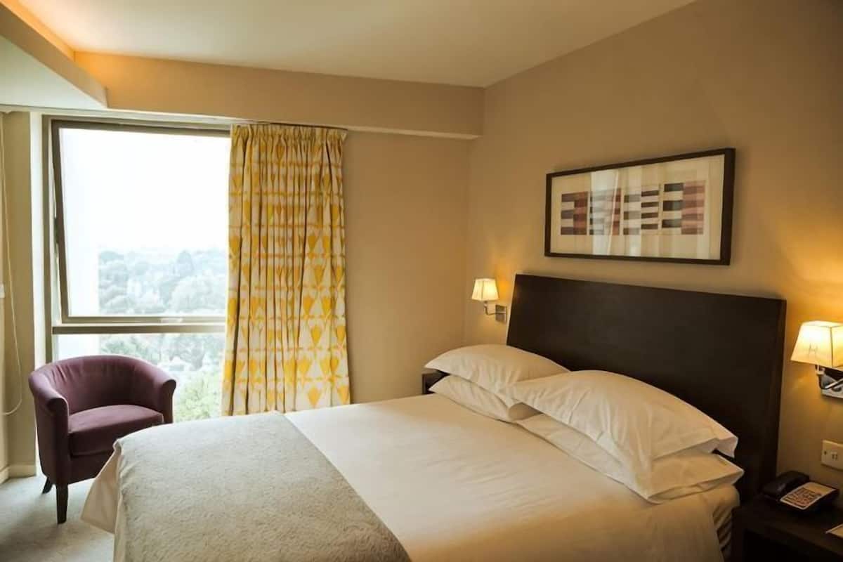 A hotel room with a bed, a chair, a window with curtains, a wall-mounted lamp, and a framed picture.