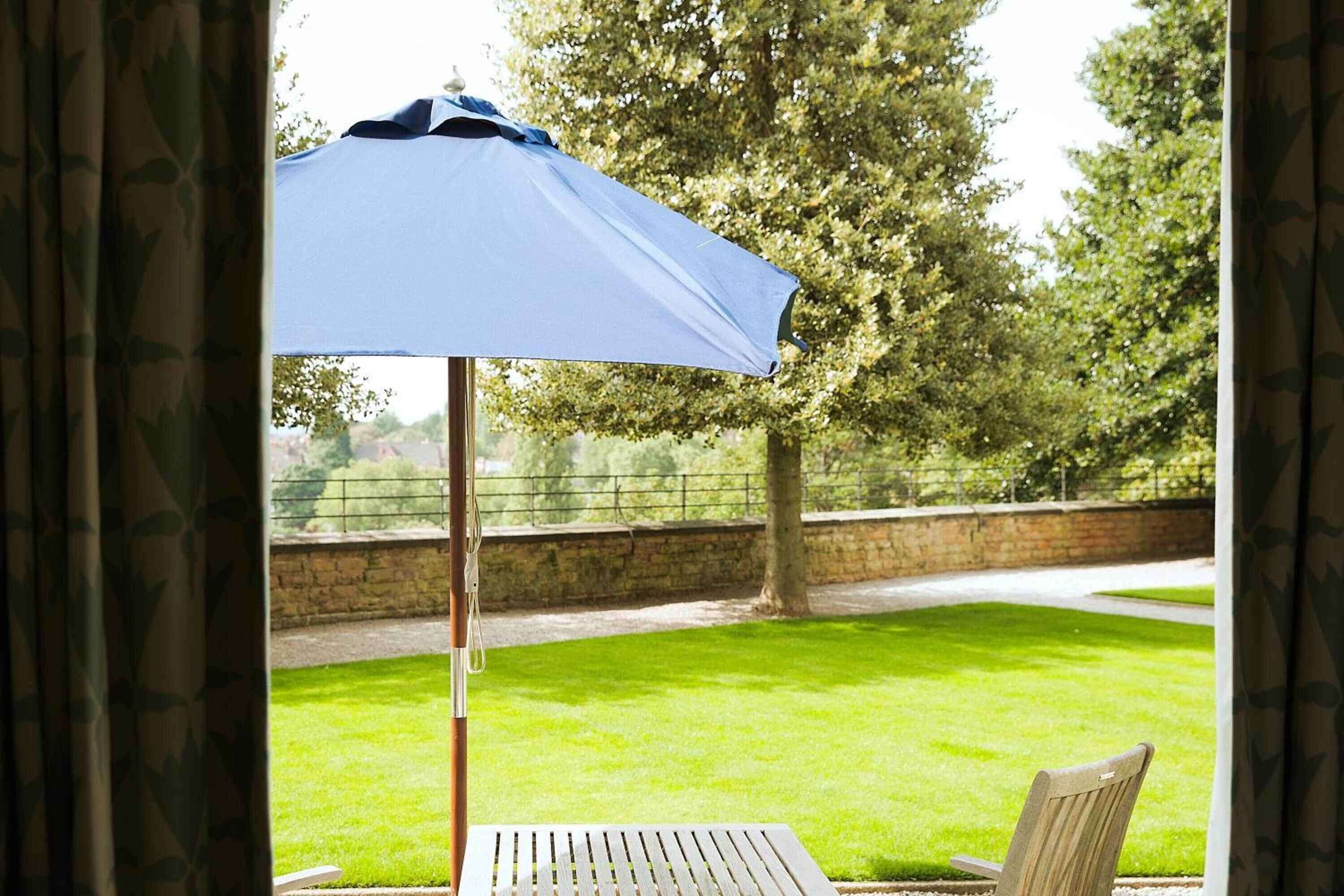 A patio with a table and a blue umbrella, overlooking a grassy area with trees and a stone wall.
