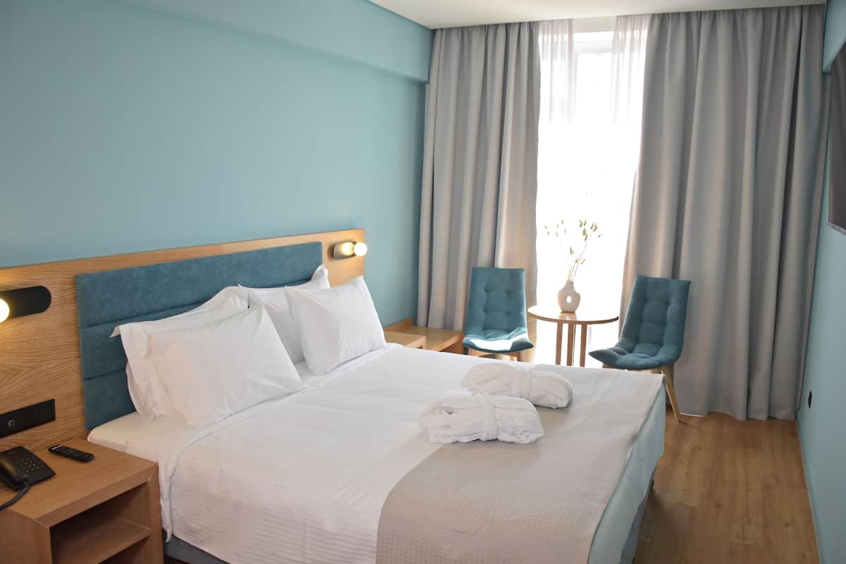 Une chambre d’hôtel avec un grand lit, une table de chevet en bois avec un téléphone, une petite table avec une chaise et une fenêtre avec des rideaux.