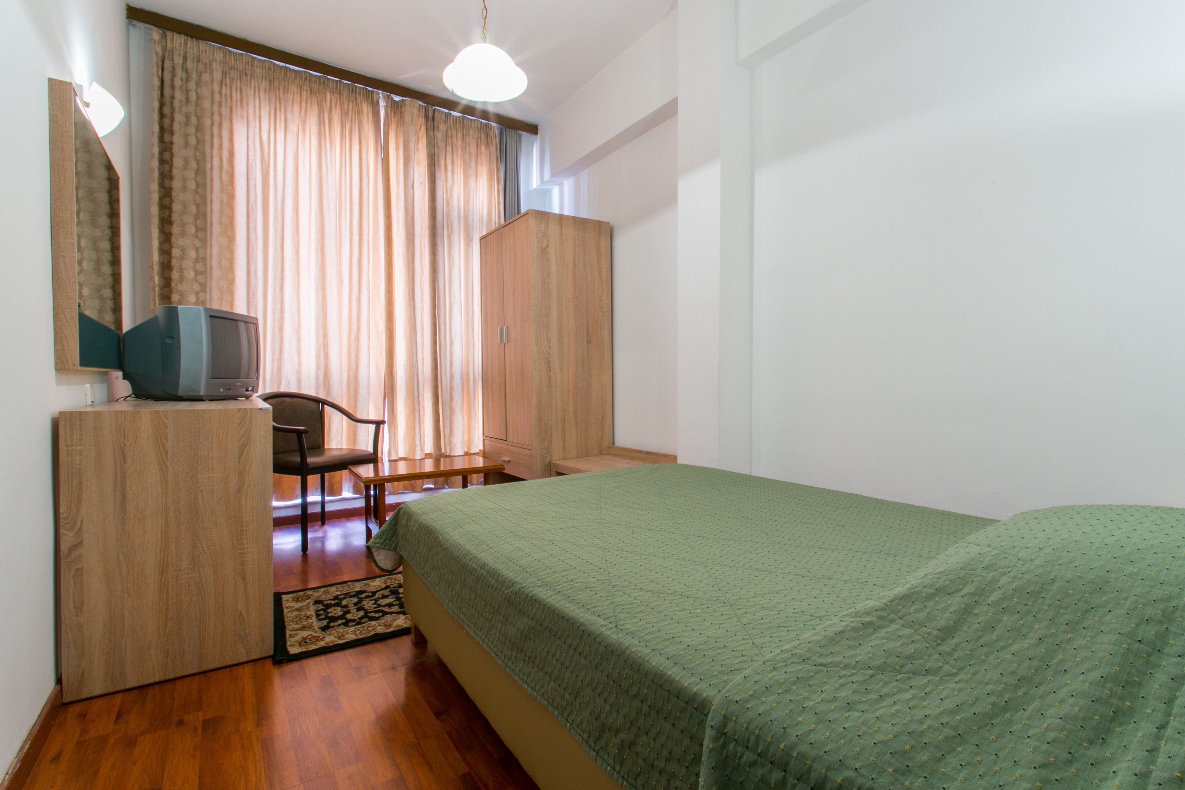 A bedroom with a bed, a wooden wardrobe, a television on a stand, a chair, and a desk.