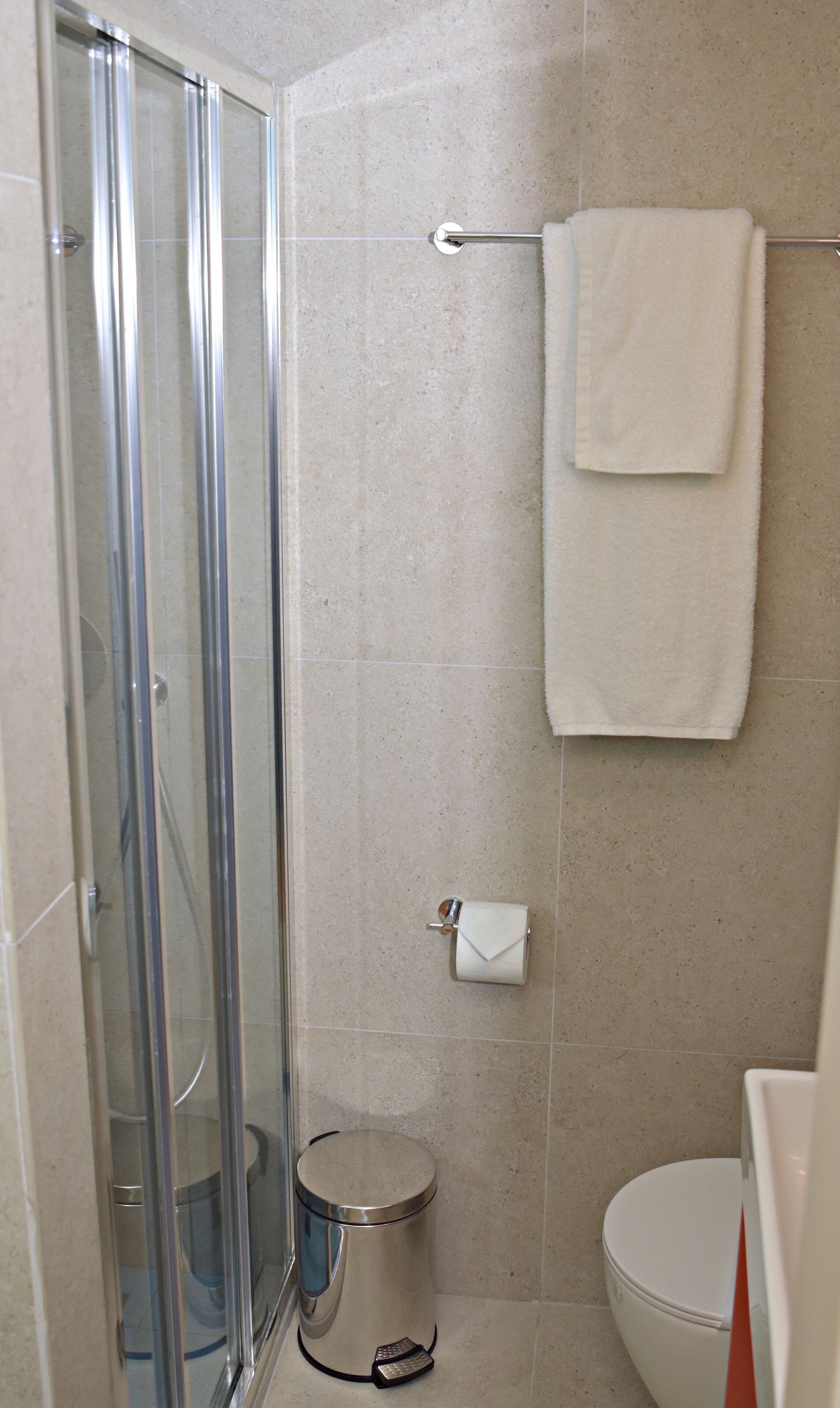 A bathroom with a glass shower enclosure, a toilet, a trash can, and a towel rack with two towels.