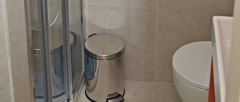 A bathroom with a glass shower enclosure, a toilet, a trash can, and a towel rack with two towels.