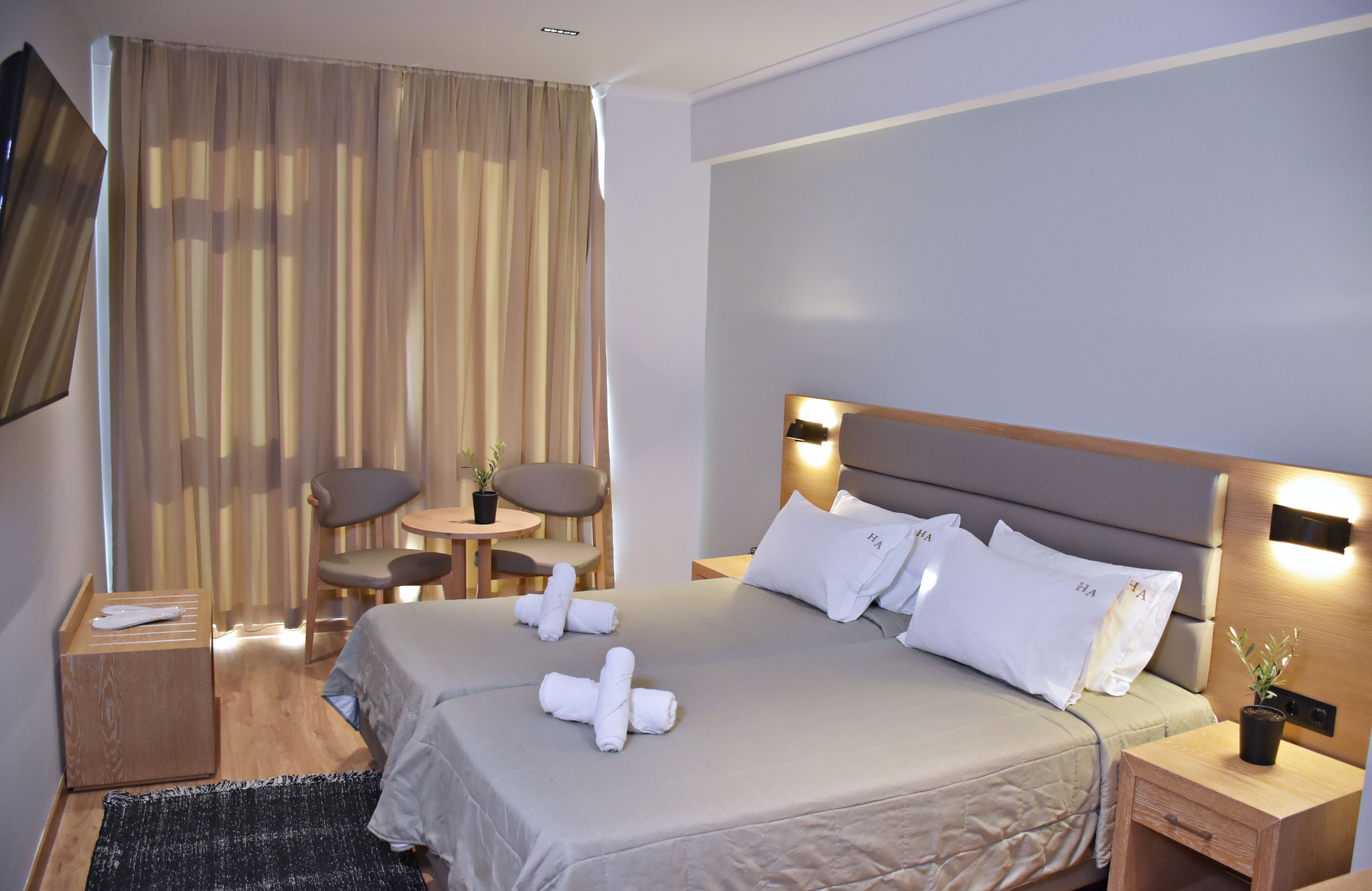 A hotel room with a large bed, two bedside tables, a small table with chairs, a TV, and a window with curtains.