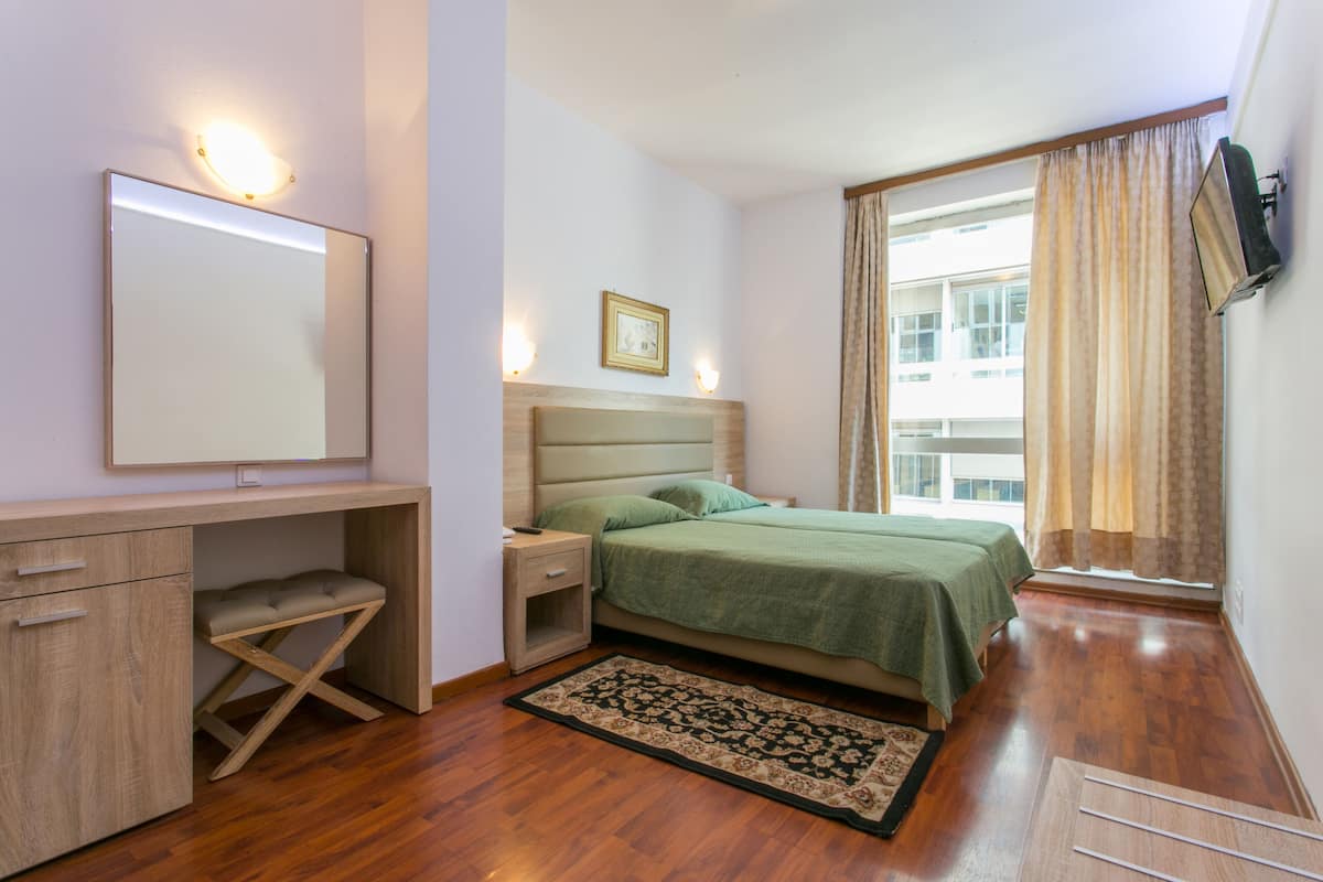 Une chambre à coucher comprenant un lit, un bureau, une chaise, une table de chevet, un miroir, un tableau accroché au mur et une fenêtre avec des rideaux.