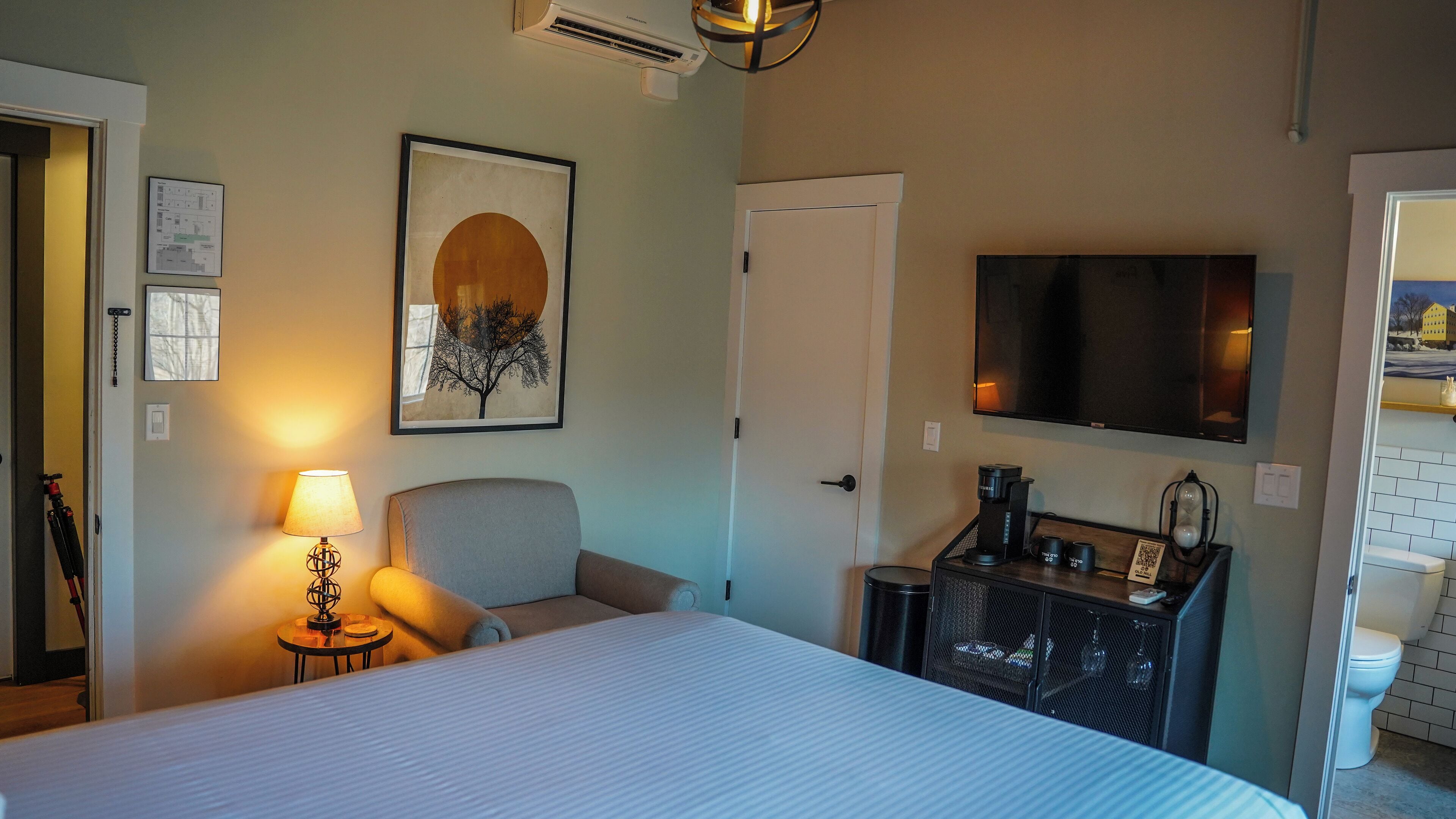 A hotel room with a bed, a chair, a desk with a coffee maker, a TV, and a framed picture on the wall.