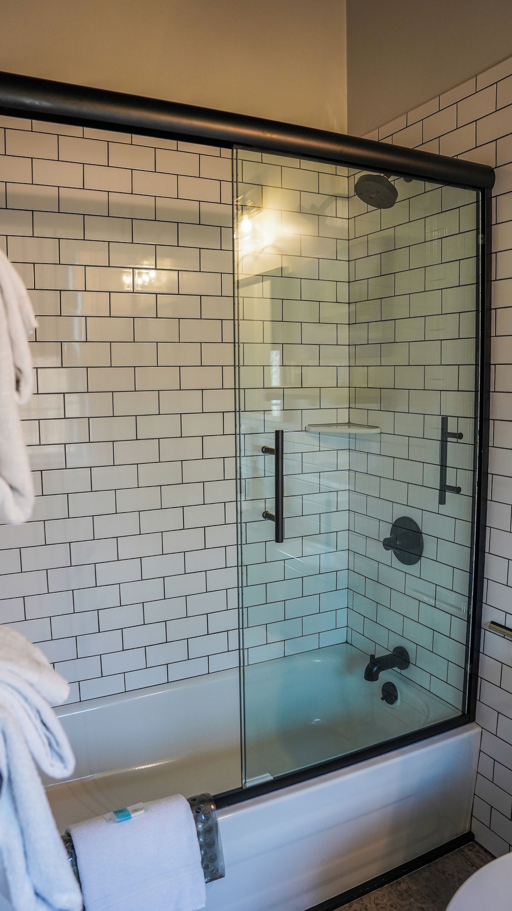 A glass-enclosed shower with a tiled wall, a black handle, and a white towel on the floor.