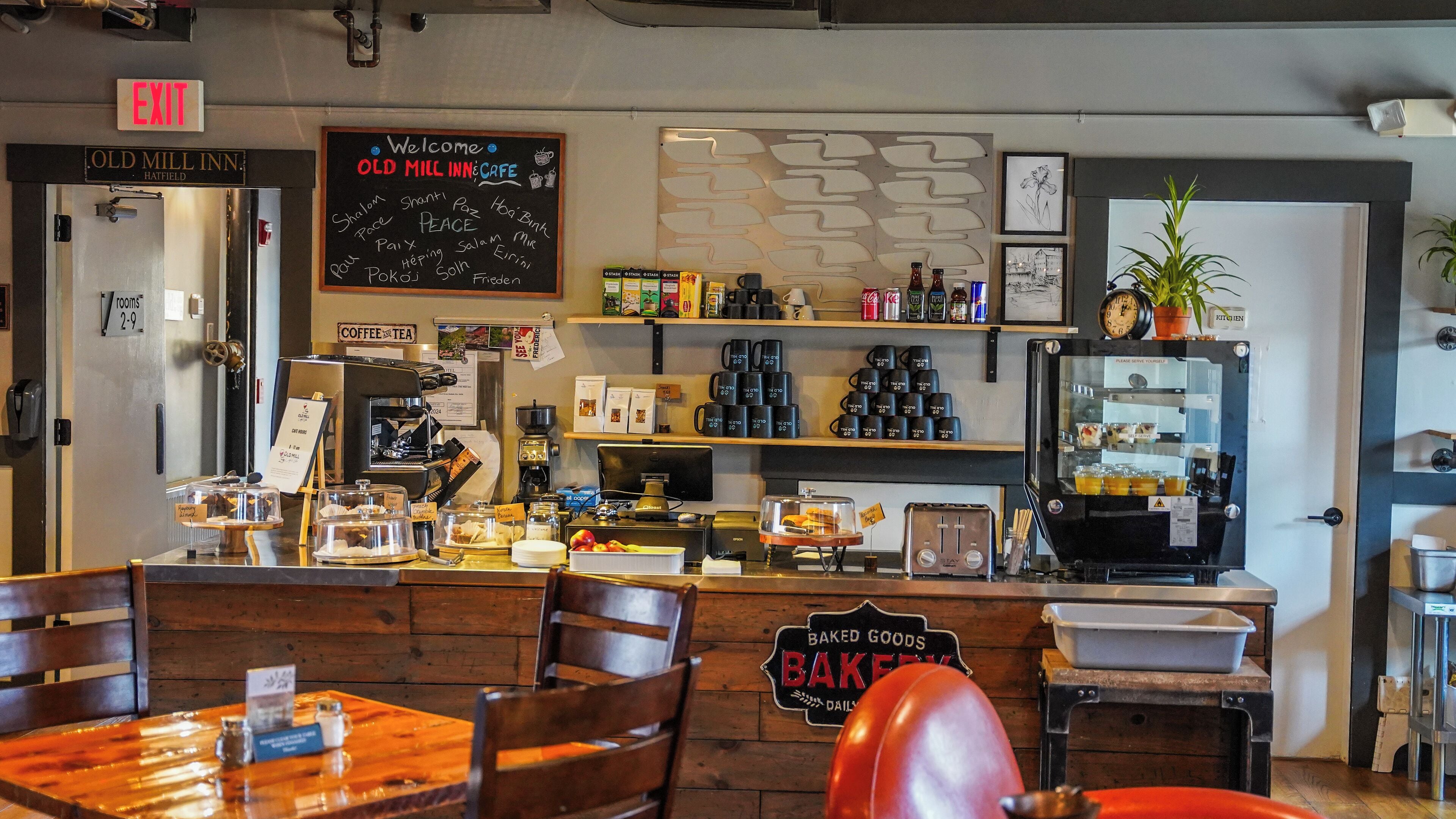 A cafe interior with a coffee machine, shelves with coffee cups, a chalkboard with 'Welcome' and 'Old Mill Inn Cafe' written on it, and a sign for 'Baked Goods'.
