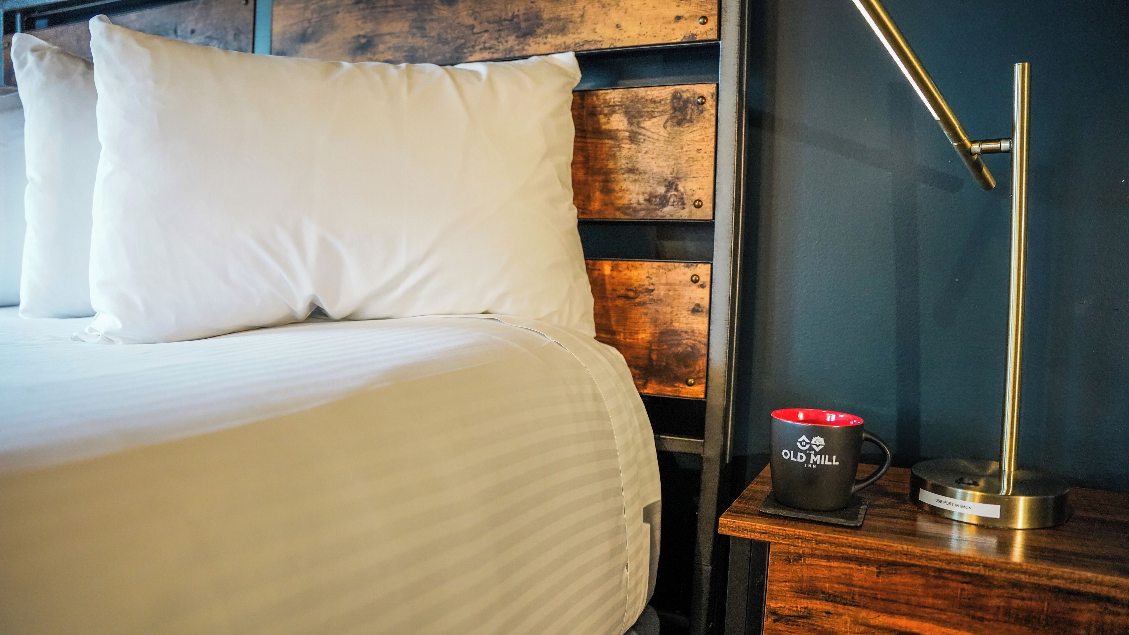 A neatly made bed with white bedding, a wooden headboard, and a black mug with white text on a wooden nightstand.