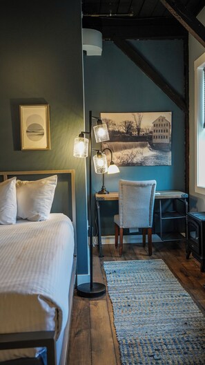A bedroom with a bed, bedside table, lamp, chair, and a painting on the wall.