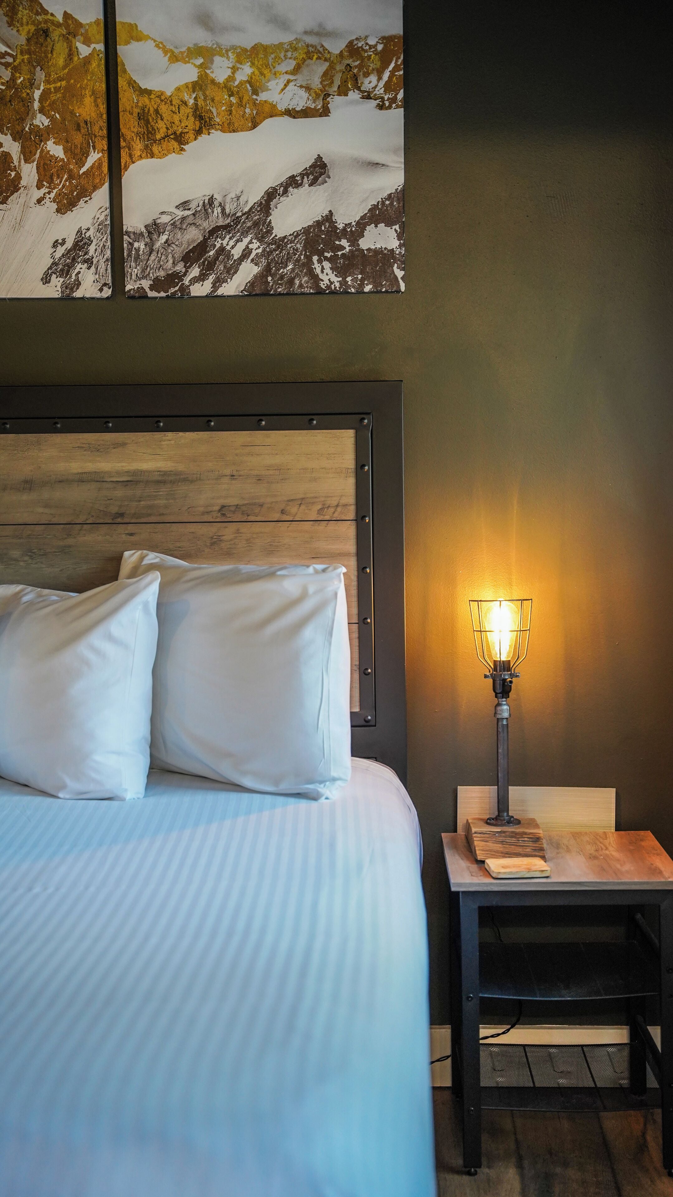A neatly made bed with white linens and pillows, a wooden headboard, a bedside table with a lamp, and a wall-mounted painting of a mountain landscape.