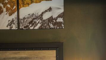 A neatly made bed with white linens and pillows, a wooden headboard, a bedside table with a lamp, and a wall-mounted painting of a mountain landscape.