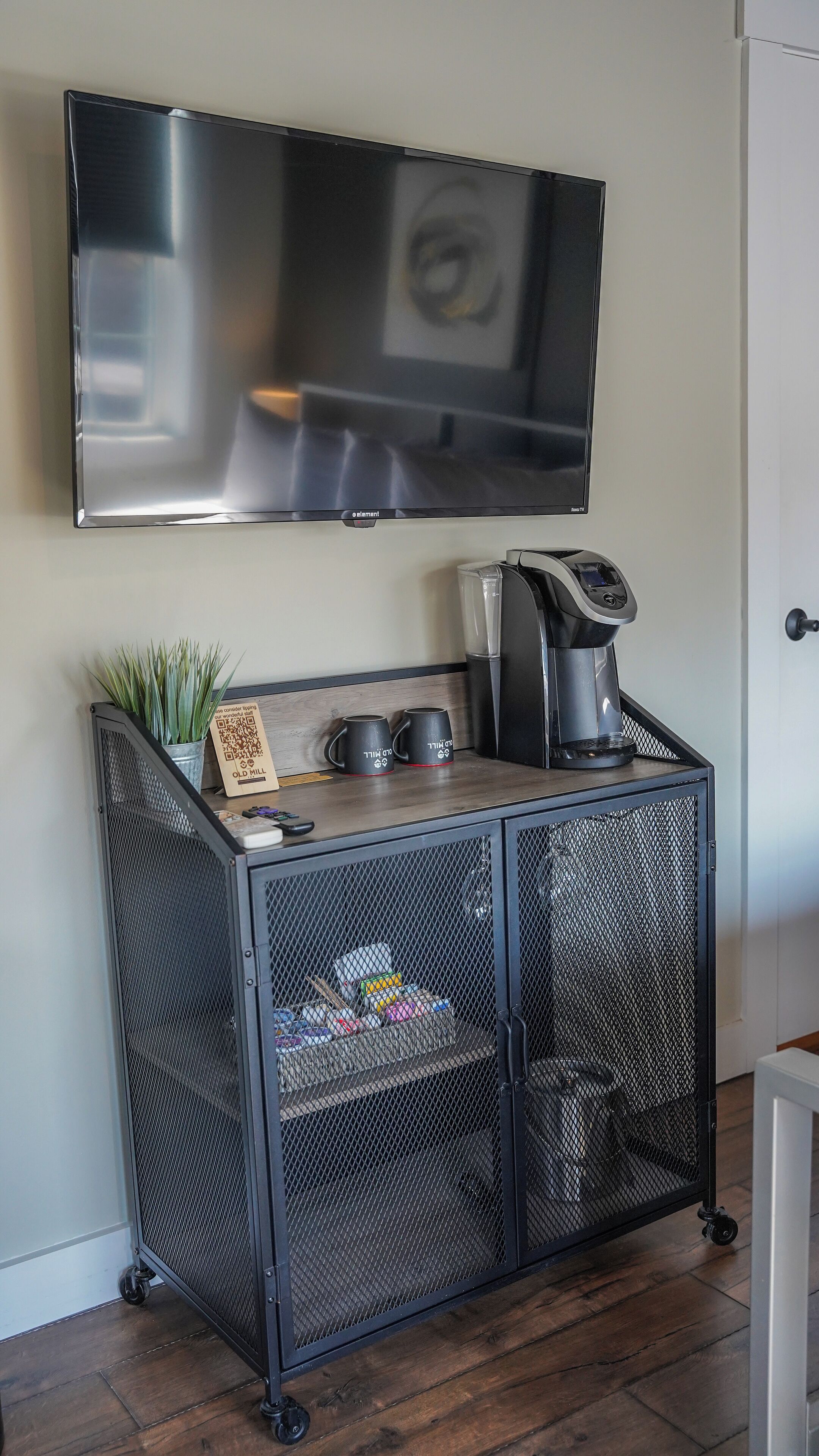 A kitchenette with a coffee maker, two mugs, a QR code sign, and a potted plant.