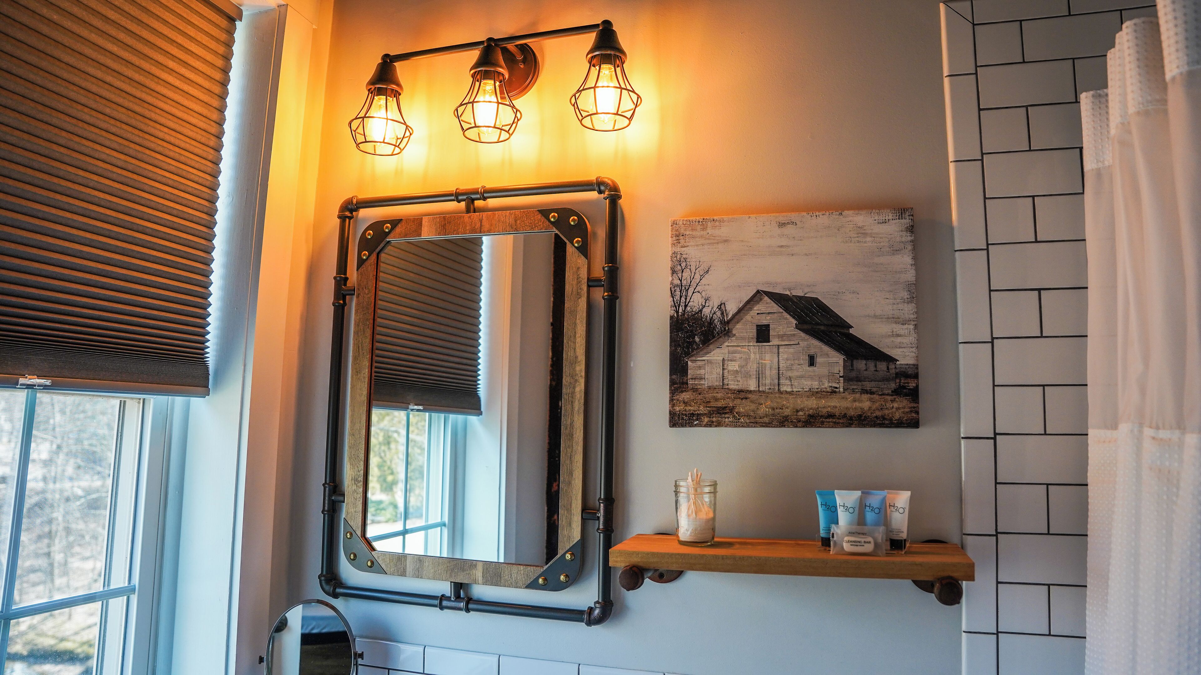 A bathroom with a wooden-framed mirror, a wall-mounted light fixture, a picture of a barn, and a shelf with toiletries.