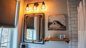 A bathroom with a wooden-framed mirror, a wall-mounted light fixture, a picture of a barn, and a shelf with toiletries. - Old Mill Inn (Hatfield)